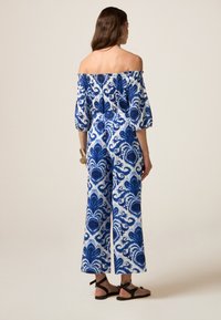 Off-the-shoulder jumpsuit van witte stof met blauwe bloemmotieven, wijde benen en een elastische taille. Gecombineerd met zwarte platte sandalen.