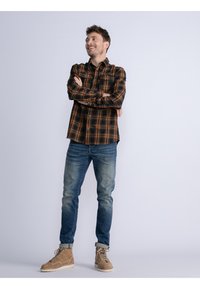 Camisa de cuadros para hombre en naranja y negro, estilo abotonado, combinada con jeans azules y botas beige. La camisa cuenta con un cuello clásico y un bolsillo con botón.