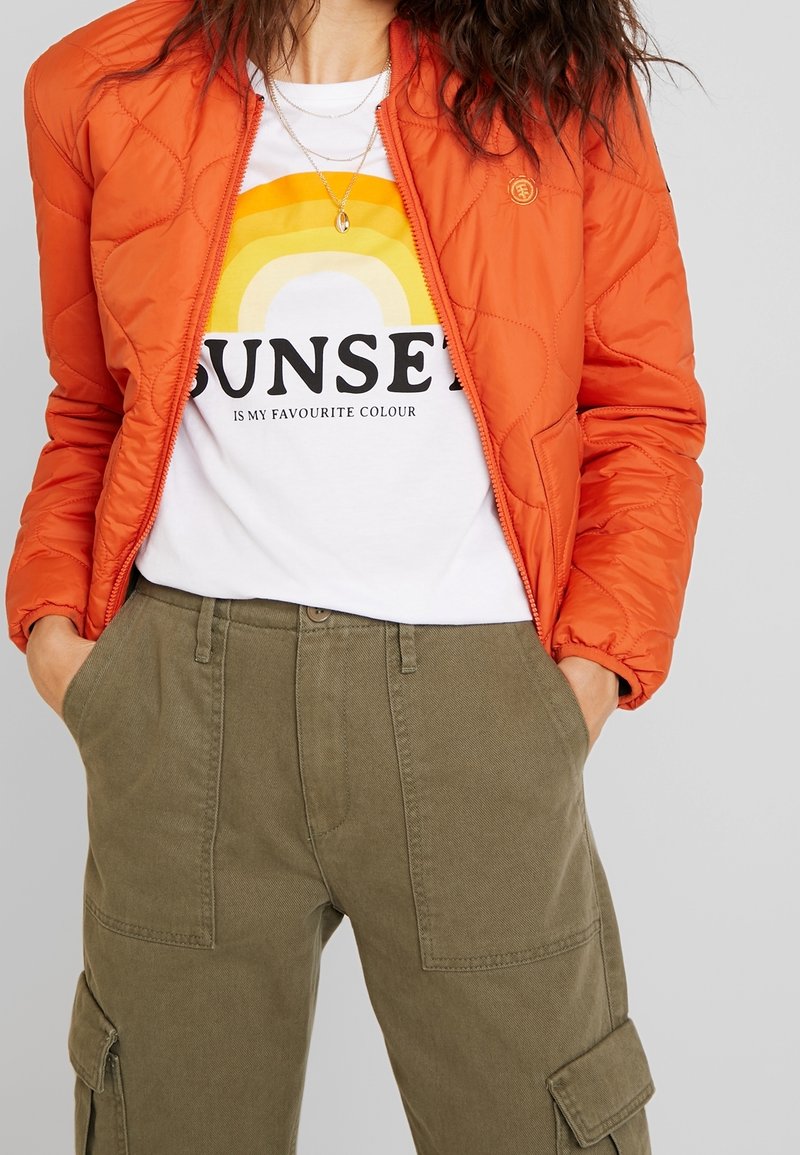 Personne portant une veste matelassée orange par-dessus un t-shirt blanc avec un motif coucher de soleil jaune et un pantalon cargo vert olive, les mains dans les poches.