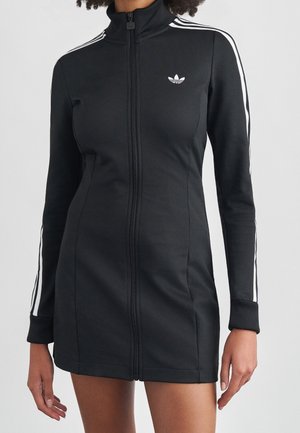 Vrouw draagt een nauwsluitende zwarte jurk met ritssluiting, witte strepen op de mouwen en het Adidas-logo op de borst, staand tegen een effen lichte achtergrond.