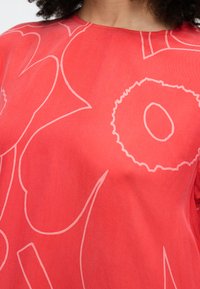 Person trägt ein korallenfarbenes Hemd mit großen, abstrakten Blumenmustern in hellrosa Linien, sichtbar sind der obere Brustbereich und der Halsausschnitt.