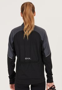 Svart sportjacka med grå axelaccenter, elastisk midja och ENX-logga på ryggen. Inkluderar figursydd design och långa ärmar.