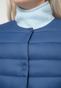 Modrá prešívaná vesta s okrúhlym výstrihom a prednými gombíkmi. Nosená cez svetlomodrý rebrovaný rolák. Hladká textura, horizontálny prešívaný dizajn.