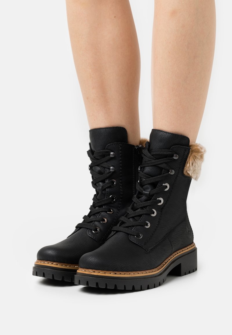 Bottes noires à lacets avec semelles épaisses et coutures marron clair, avec bordure en fourrure en haut, portées sur jambes nues sur fond blanc.