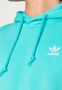 Tyrkysová mikina s kapucí a šňůrkami, s malým bílým logem Adidas. Měkká, texturovaná tkanina s klasickým střihem přes hlavu.