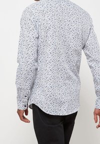 Košile s dlouhým rukávem s světle modrým květinovým vzorem na bílém pozadí, vyrobená z měkké látky, s prohnut�ým lemem a manžetami na knoflíky.