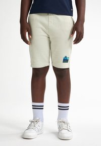 Lichtbeige, knie-lange shorts van zachte stof met een opgebogen zoom. Heeft een palmboomgrafiek en wordt gedragen met witte schoenen en gestreepte sokken.