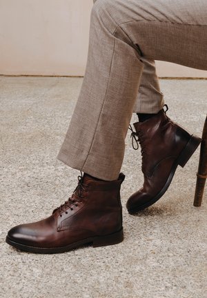 Brown leather ankle boots with a rounded toe, lace-up design, and a subtle textured finish, paired with light checkered trousers.