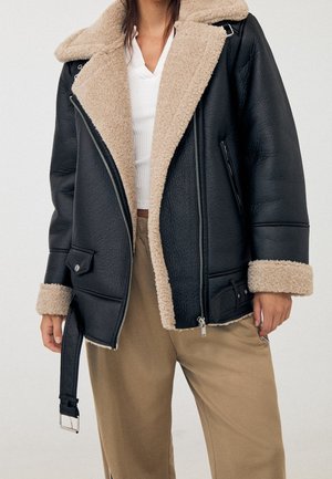 Persoon die een zwarte leren jas met beige shearling voering draagt, een wit topje en beige broek, staande tegen een effen lichte achtergrond.