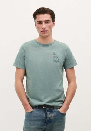 Lichtgroene katoenen t-shirt met ronde hals, korte mouwen en bedrukte tekst op de linkerborst. Gepaard met blauwe spijkerbroek.
