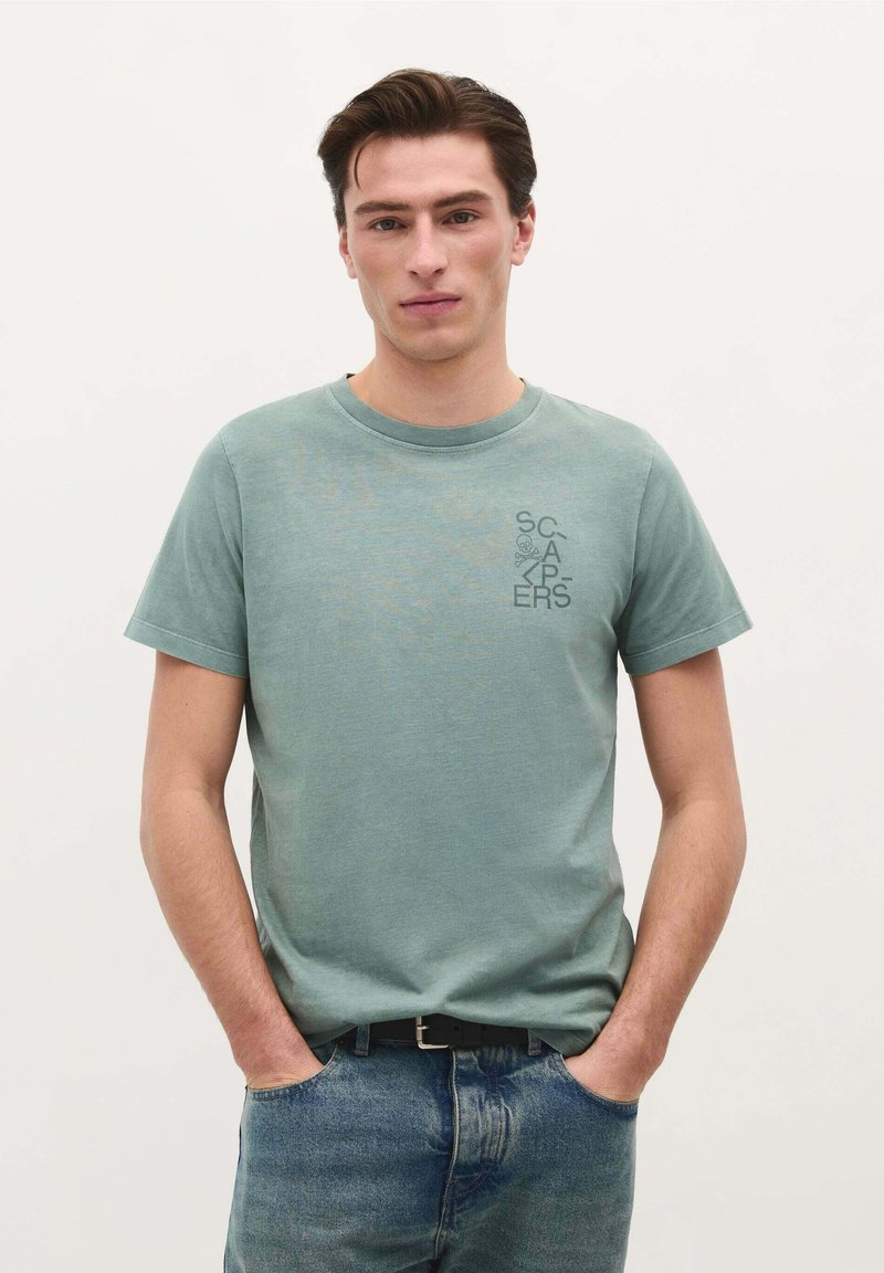 Hellgrünes Baumwoll-T-Shirt mit Rundhalsausschnitt, kurzen Ärmeln und bedrucktem Text auf der linken Brust. Kombination mit blauen Jeans.
