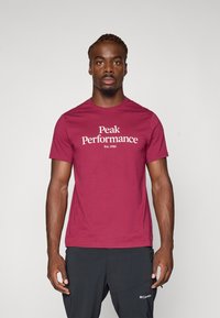 Bordeauxfarbenes Baumwoll-T-Shirt mit "Peak Performance" Logo in weißem Text, Rundhalsausschnitt, kurzen Ärmeln und einer taillierten Silhouette.