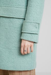Cappotto verde menta con texture, colletto ampio e polsini oversize. Mano con tatuaggio a forma di cuore visibile, sovrapposta a tessuto a quadri.