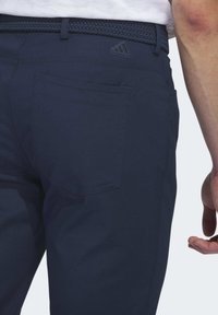 Marineblauwe broek gemaakt van een glad materiaal, met een geweven riem en een achterzak met een tonale logodetail. Eenvoudig, op maat gemaakt ontwerp.