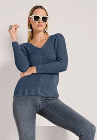 Blauwe gebreide trui met een V-hals, textuurpatroon, geribde manchetten en onderrand. Gepaard met grijze jeans en oversized witte zonnebril.