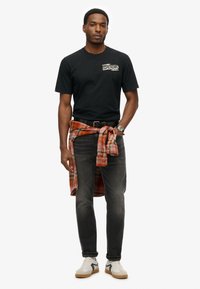 T-shirt nera in cotone con logo, jeans grigi, camicia di flanella a quadri legata in vita e sneakers bianche con suola marrone e accenti grigi.