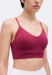 Γυναίκα φοράει ροζ αθλητικό σουτιέν Under Armour με λεπτές τιράντες και άσπρο κολάν, εμφανίζεται από τους ώμους μέχρι τη μέση σε απλό φόντο.