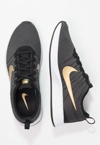 Zapatillas deportivas negras con una parte superior de malla texturizada, emblema dorado de Nike, cordones negros y una entresuela blanca. Presenta una lengüeta en el talón.