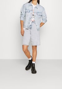 Giacca di jeans azzurro chiaro, t-shirt grafica, pantaloncini chiari e stivaletti neri alla caviglia con calzini bianchi. Outfit casual a strati con texture variegate.