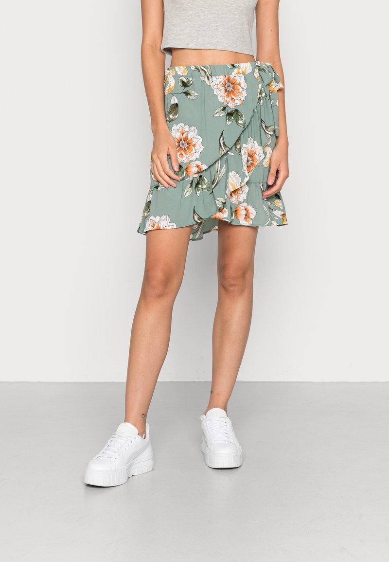 Femme portant une jupe portefeuille florale verte avec des volants, associée à un crop top gris et des baskets à plateforme blanches, debout sur un sol gris.