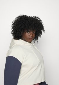 Hoodie accorciato con corpo crema, maniche blu navy e cordoncino. Texture liscia. Design semplice, adatto per un abbigliamento casual.