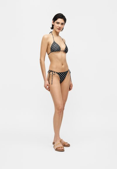 Γυναίκα όρθια, φοράει τρίγωνο μαγιό με ασπρόμαυρα σχέδια και δέσιμο, καθώς και πλεκτά σανδάλια σε απλό λευκό φόντο.