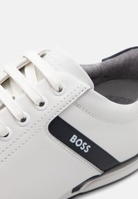 Baskets en cuir blanc avec un design à lacets, des œillets ronds, une bande d'accent bleu marine et une doublure intérieure grise texturée. Logo "BOSS" affiché.