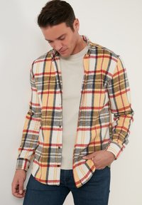Flanellen shirt met een gele, rode en grijze ruitpatroon; knoopsluiting, lange mouwen en zachte textuur. Gelaagd over een lichtbeige shirt.