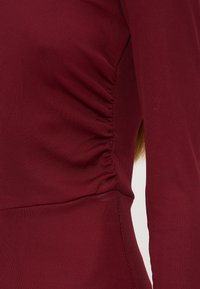 Gros plan d'un haut côtelé bordeaux à manches longues montrant un détail de tissu froncé sur le côté près de la taille.