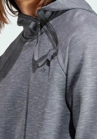 Närbild av en person som bär en grå zip-up hoodie med en uppfälld krage och en liten, subtil logotyp på bröstet.