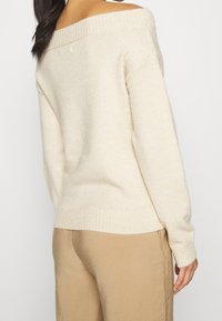 Krämfärgad tröja med off-shoulder-design, ribbat halsringning och mudd, med en mjuk textur och avslappnad passform. Beigefärgade byxor synliga.