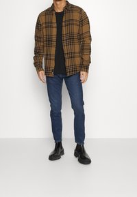 Ruitjes flanellen shirt in bruin en zwart, gedragen over een zwart t-shirt, gecombineerd met blauwe jeans en zwarte chelsea boots. Focus op de textuur en het patroon van het shirt.