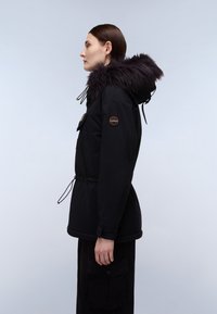 Sort isoleret jakke med faux fur hætte, har snoretræk i taljen, sidelommer og et patch på venstre ærme. Glat tekstur.