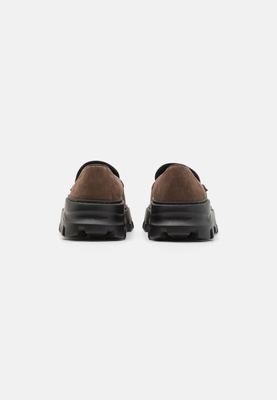 Braune Wildleder-Slipper mit einer massiven schwarzen Sohle, im runden Design und minimalistischem Stil auf einem schlichten weißen Hintergrund.