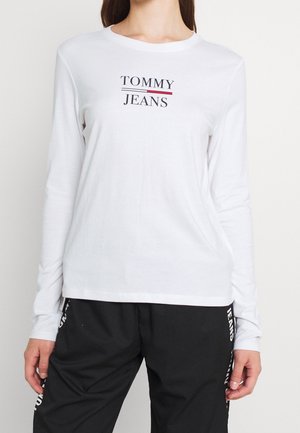 T-shirt à manches longues blanc avec "TOMMY" en bleu marine et "JEANS" en rouge. Tissu doux ; encolure ronde et coupe décontractée. Associé à un pantalon noir.