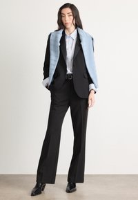 Zwart op maat gemaakt pak met een getailleerde blazer, een lichtblauwe gebreide omslag en een lichtblauw gestreept shirt; gecombineerd met zwarte enkellaarsjes met hak.