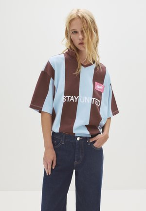 T-shirt en blocs de couleur avec des rayures verticales bleues et brunes, présentant un col en V et un texte blanc "RESTEZ UNIS", accompagné d'une petite étiquette de logo rose.