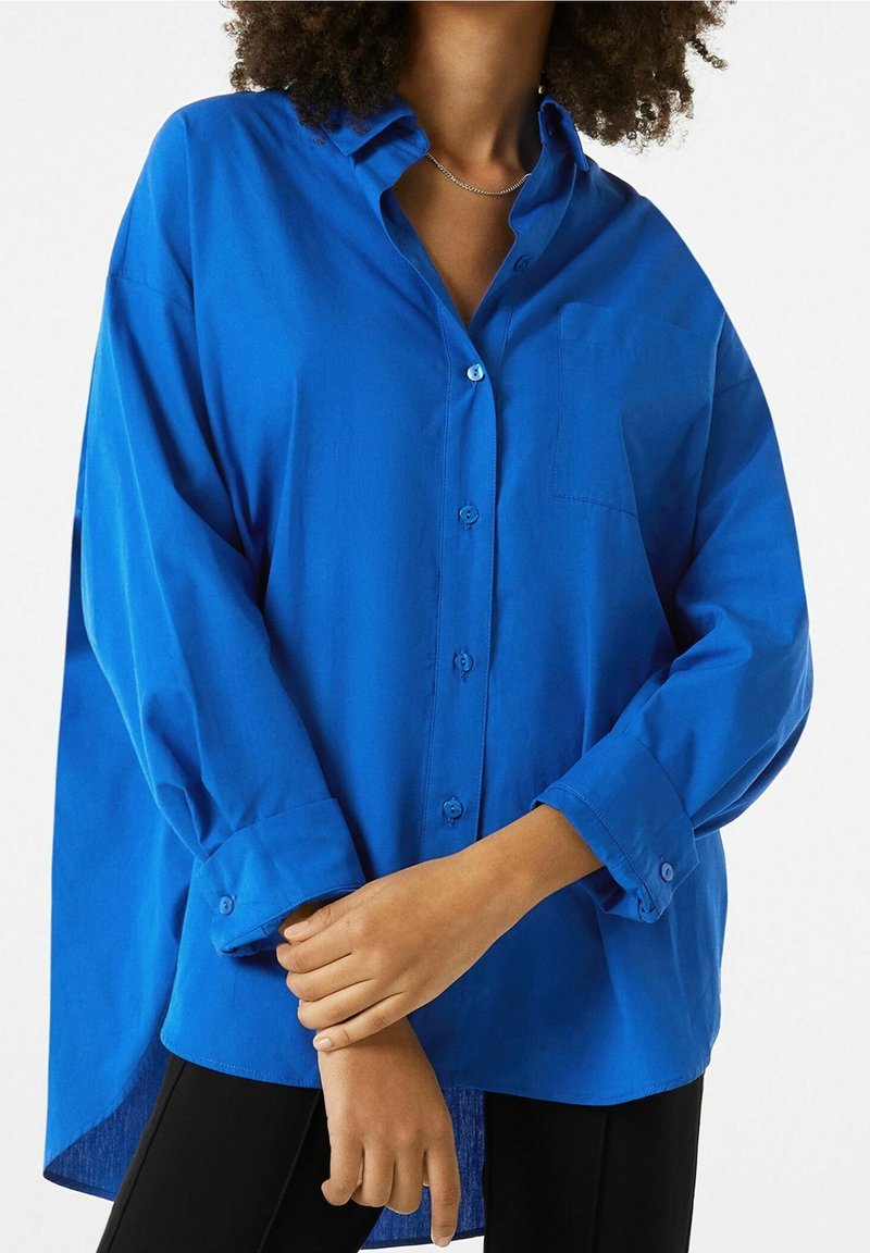 Camisa azul de manga larga con botones y cuello, un bolsillo en el lado izquierdo del pecho y un dobladillo trasero ligeramente más largo. Textura de tela suave.