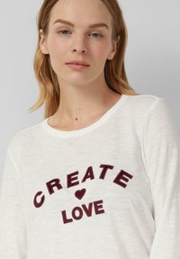 T-shirt léger à manches longues blanc avec le texte brodé en maroon "CREATE ♥ LOVE" dans une disposition courbée. Texture douce et design à col rond.
