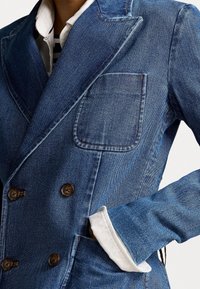 Modrý denimový blazer s dvouřadým zapínáním, velkou přední kapsou a hnědými knoflíky. Obsahuje bílý detail na rukávech.