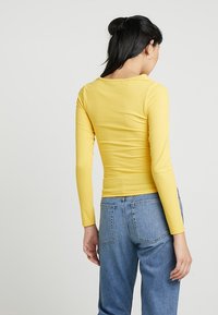 Žlutý mikinový top s dlouhými rukávy a upnutým střihem, který má široký výstřih. Nosený s modrými džíny s vysokým pasem.