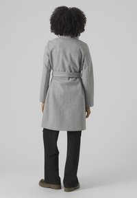 Manteau en laine gris à taille ceinturée, à manches longues et coupe droite, associé à un pantalon large noir et des chaussures vertes.