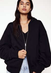 Zwarte rits hoodie met een ontspannen pasvorm, grote capuchon en ribgebreide manchetten. Kenmerkt zich door een zilveren rits en zachte, gestructureerde stof.