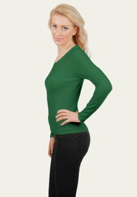 Haut vert à manches longues avec un col en V, tissu texturé, design ajusté, associé à un jean noir, modèle posé de profil avec une main sur la hanche.