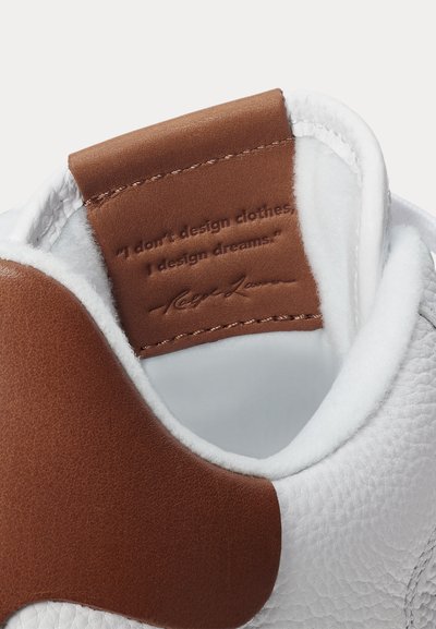 Adidași albi cu detaliu din piele maro pe care este text embosat: „Nu proiectez haine, proiectez vise.” și o semnătură.