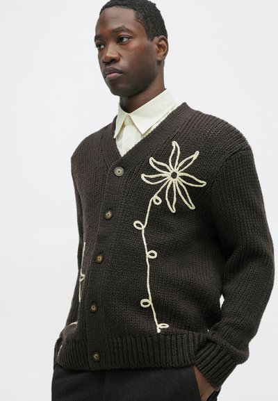 Barna kötött kardigán V-nyakkal, melyen krémszínű virágos hímzés található elöl, valamint barna gombos részletek. Puha tapintás.