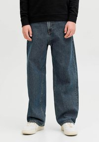 Személy laza kék farmert, fehér tornacipőt és fekete hosszú ujjú felsőt viselve, sima világos háttér előtt áll.