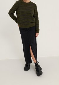 Olivgrüner Pullover mit strukturiertem Zickzackmuster und langem schwarzen Denimrock mit einem oberschenkelhohen Schlitz, kombiniert mit klobigen schwarzen Schnürstiefeln.