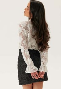 Vit spetsblus med blommönster och utsvängda ärmar, parat med en svart texturerad minikjol. Outfitten är figurnära och feminin.