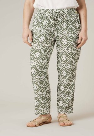 Vue de la taille vers le bas d'une personne portant un pantalon à motif blanc et vert olive avec une taille élastique et des sandales plates dorées à fines lanières.